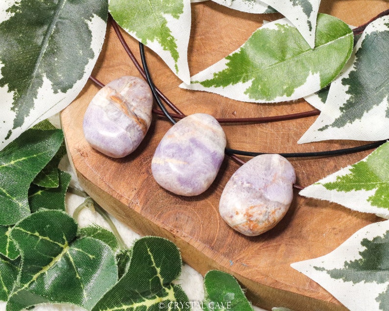 Puede incluir: Tres piedras pulidas de forma ovalada, en tonos lavanda, beige y marr&oacute;n, ensartadas en cordones negros. Se exhiben sobre una superficie de madera, rodeadas de acentos de hojas verdes y blancas. La imagen resalta las texturas y colores naturales de las piedras.