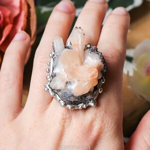 May include: A silver ring with a large, pink and white crystal cluster set in the center. The crystal cluster is surrounded by a silver band with small beads.