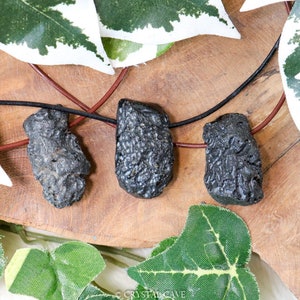 Könnte beinhalten: Drei schwarze Vulkangestein-Anhänger an einem braunen Lederband. Die Anhänger sind rau und strukturiert, mit einer dunklen, matten Oberfläche.