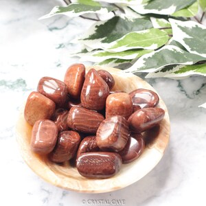 May include: A bowl of tumbled red jasper stones. The stones are smooth and polished, with a reddish-brown color. The bowl is made of a light-colored material.