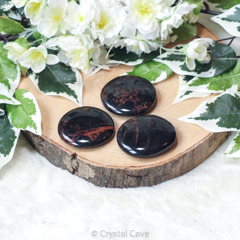 May include: Three smooth, polished black and red mahogany obsidian palm stones. The stones are arranged on a wooden slice.