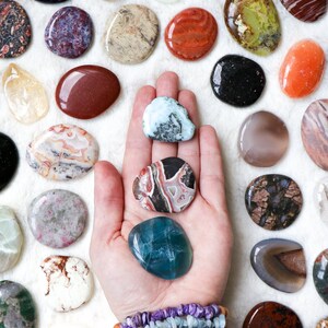 May include: A variety of polished, smooth, oval-shaped stones in different colors and patterns, including green, brown, red, white, and black. The stones are arranged in a circle around a hand holding three stones: a light blue stone, a red and black stone, and a green and white stone. The hand is wearing a bracelet with multiple strands of beads in various colors.