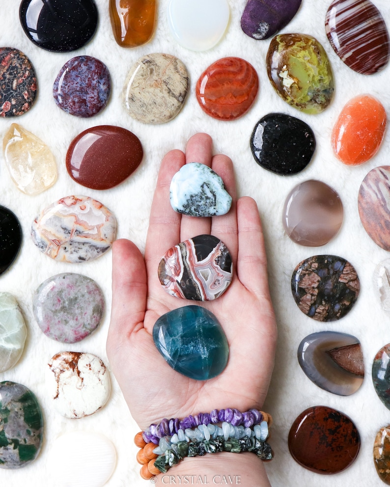 May include: A variety of polished, smooth, oval-shaped stones in different colors and patterns, including green, brown, red, white, and black. The stones are arranged in a circle around a hand holding three stones: a light blue stone, a red and black stone, and a green and white stone. The hand is wearing a bracelet with multiple strands of beads in various colors.