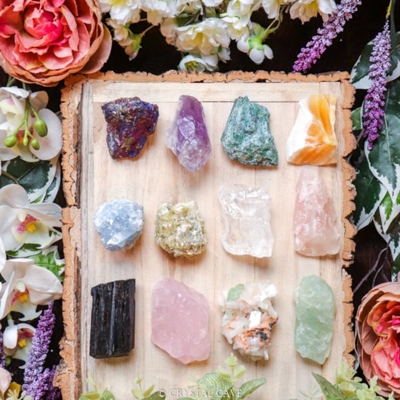May include: A collection of 12 different types of crystals, including amethyst, rose quartz, clear quartz, and green aventurine. The crystals are arranged on a wooden board with a light brown background. The crystals are all natural and have a variety of shapes and sizes.
