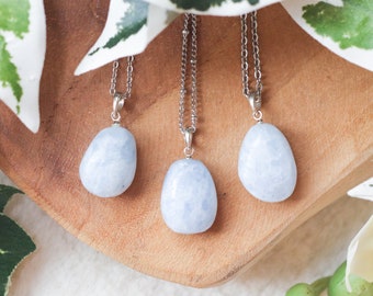 May include: Three silver necklaces with light blue, oval-shaped gemstone pendants. The necklaces are displayed on a wooden surface with green leaves in the background.