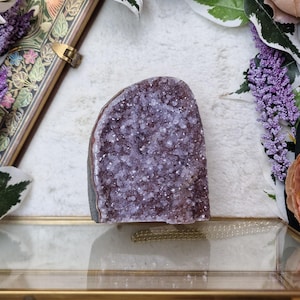 May include: A large, purple amethyst geode with a natural, rough exterior and a smooth, crystal-lined interior. The geode is displayed on a glass shelf with a gold frame.