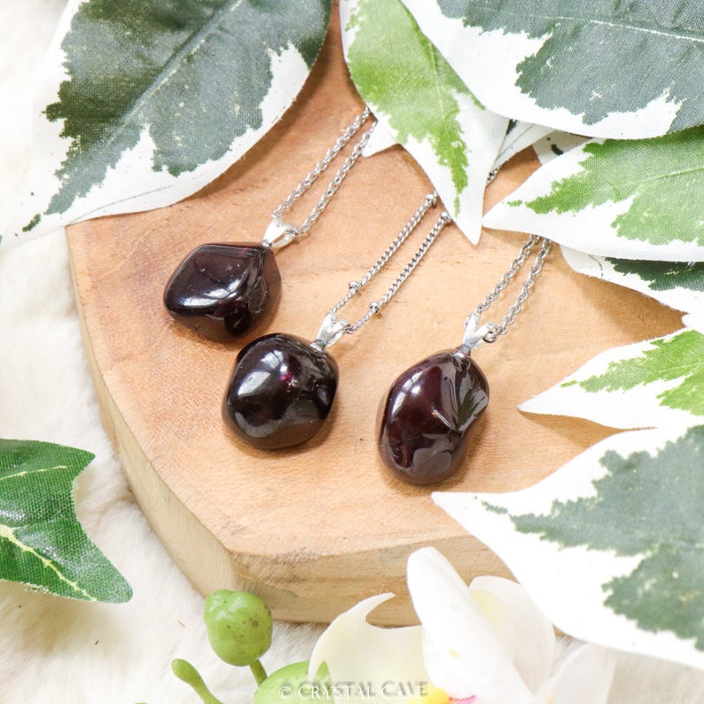 May include: Three silver necklaces with dark red gemstone pendants on a wooden surface. The necklaces are on a background of green and white leaves.