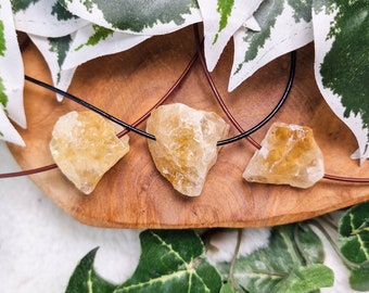 Puede incluir: Tres colgantes de cristal de citrino en bruto, naturales, de color amarillo-marrón, en cordones negros. Los cristales están sobre una superficie de madera con hojas verdes en el fondo.