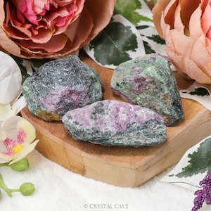 Op de afbeelding: Drie ruwe, groene en roze robijnzoisietkristallen op een houten oppervlak. De kristallen hebben een natuurlijke, ruwe uitstraling.