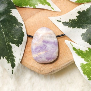 Puede incluir: Un colgante de amatista ovalado pulido sobre un cord&oacute;n negro. La piedra presenta una mezcla de tonos lavanda y blancos. El colgante descansa sobre una peque&ntilde;a superficie de madera, rodeado de hojas verdes y blancas. El texto "CRYSTAL CAVE" est&aacute; en la esquina inferior derecha.