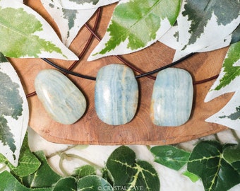 May include: Three light blue, oval-shaped, polished stones on a brown wooden surface. The stones are strung on a black cord and are surrounded by green and white leaves.