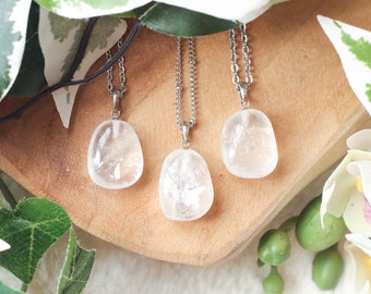 May include: Three clear quartz crystal pendants on silver chains. The pendants are smooth and oval shaped. The chains are hanging from a wooden surface.