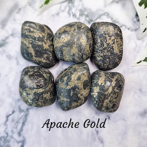 May include: Six Apache Gold tumbled stones. The stones are a dark gray color with flecks of gold throughout. The stones are smooth and rounded. The text "Apache Gold" is written in a black script font.