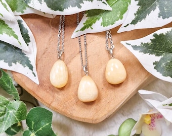 Puede incluir: Tres collares de plata con colgantes de cristal amarillo en forma de lágrima. Los collares se muestran sobre una superficie de madera con hojas verdes en el fondo.