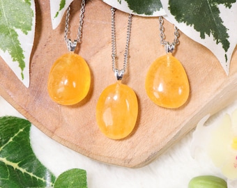 Puede incluir: Tres colgantes de piedra de cornalina amarilla en cadenas de plata. Las piedras son lisas y de forma ovalada. Los colgantes se muestran sobre una superficie de madera.