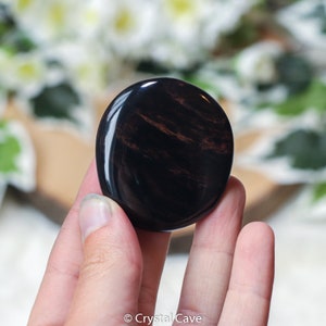 May include: A smooth, polished black obsidian palm stone with red and brown striations. The stone is held in a hand, with the fingers visible.