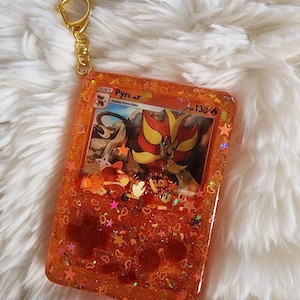 Pode incluir: Chaveiro de resina laranja e dourado com glitter, apresentando um cartão Pokémon com uma imagem de um Pyroar, um Pokémon do tipo fogo. O texto do cartão diz "Pyroar, Evolui de Litleo, N.º 130".