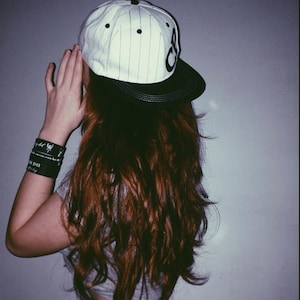 May include: A woman with long, brown hair wearing a white and black baseball cap with a black brim. The cap has a white stripe pattern and black lettering.