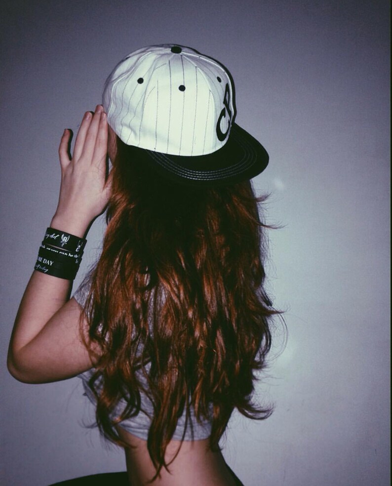 May include: A woman with long, brown hair wearing a white and black baseball cap with a black brim. The cap has a white stripe pattern and black lettering.