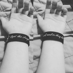May include: Two black wristbands with white text. The left wristband says "Live" the Angel. The right wristband says "aid and so they did."