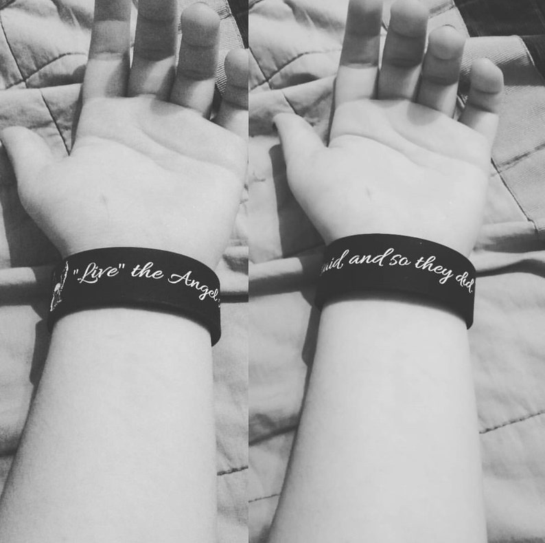 May include: Two black wristbands with white text. The left wristband says "Live" the Angel. The right wristband says "aid and so they did."