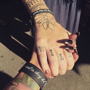 May include: Two people holding hands, both with tattoos on their fingers. The person on the left has a black wristband with the text "and so they did". The person on the right has a black wristband with the text "can be the book".