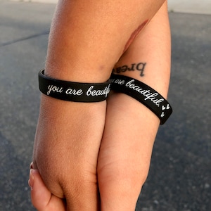 May include: Two black silicone bracelets with white text. The bracelet on the left says "you are beautiful". The bracelet on the right says "you are beautiful" with two small hearts.