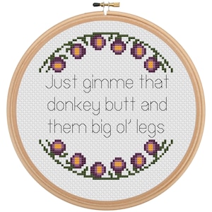 May include: Cross-stitch embroidery in a wooden hoop. The design features the text "Just gimme that donkey butt and them big ol' legs" in black, with a floral border of purple and yellow flowers. The background is white.
