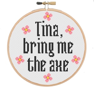 Könnte beinhalten: Ein weißer Stickrahmen mit dem Text "Tina, bring me the axe" in schwarzen Buchstaben. Um den Text herum sind rosa Blumen gestickt.