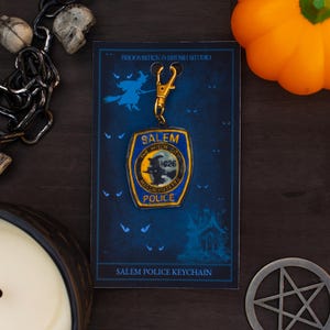 May include: A Salem Police keychain with a gold-coloured clasp and a blue and gold badge. The badge features a witch silhouette and the text "Salem Police 1626". The keychain is displayed on a card with a witch graphic and the text "Salem Police Keychain".