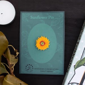 May include: A yellow sunflower pin attached to a rectangular green card with the text Sunflower Pin and Broomstick & Brush Studio printed on it. The pin features a bright yellow flower with a small green centre, displayed on a dark surface.