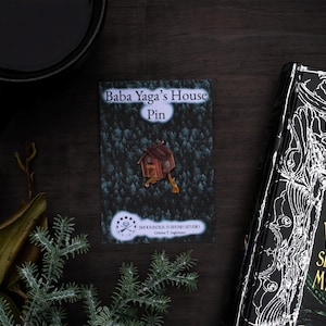 Puede incluir: Un pin con el texto "Baba Yaga's House Pin" que presenta una pequeña casa de madera sobre un fondo de bosque. El pin se muestra sobre una superficie de madera oscura junto a un libro titulado "Witches, Wizards, Seers & Healers, Myths & Tales".