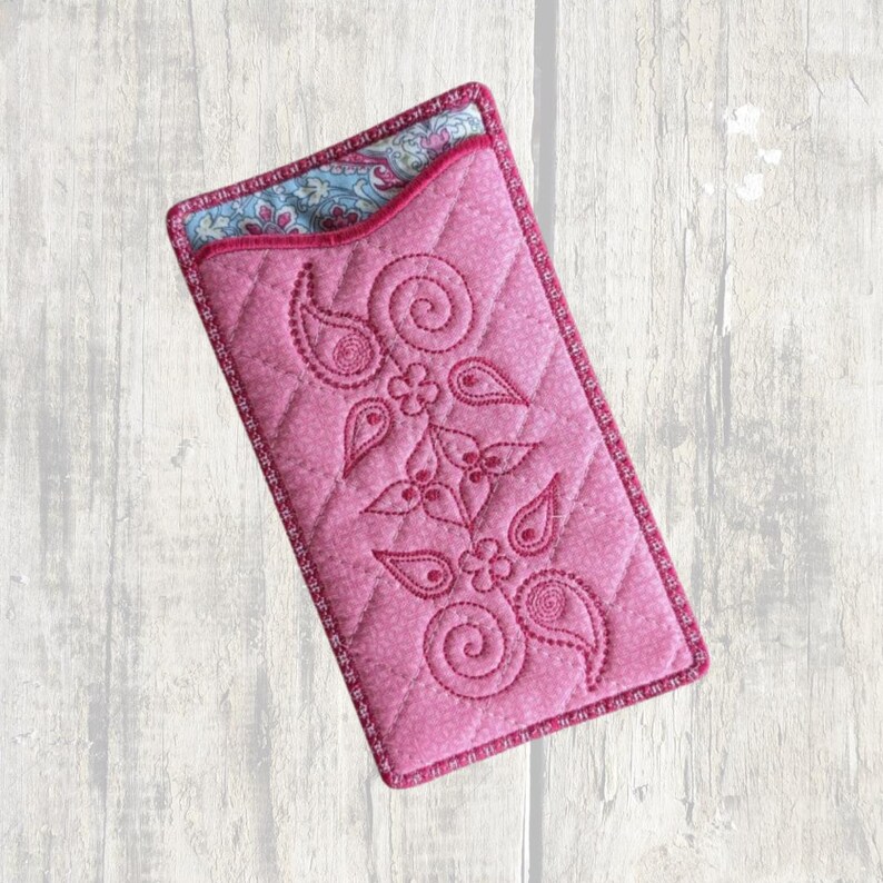 May include: A rectangular pink quilted fabric pouch featuring a stitched floral and paisley pattern on the front. The top opening reveals a contrasting interior lining with a blue, pink, and white floral print. The edges are finished with pink trim.