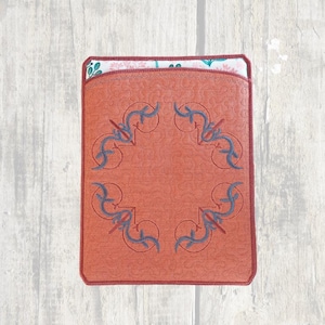 May include: A red leather pouch with a decorative blue embroidered design. The pouch has a flap with a white and pink floral pattern.