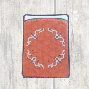 May include: A rectangular, quilted, orange case with a decorative white and gray floral design. The case has a dark blue border and a white interior. The case is sitting on a light wood surface.