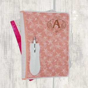 May include: A coral-colored journal cover with a starfish pattern and the letter "A" embroidered in a decorative frame. A white pen holder is attached to the cover, holding a silver pen. The journal has a white border and a pink interior.