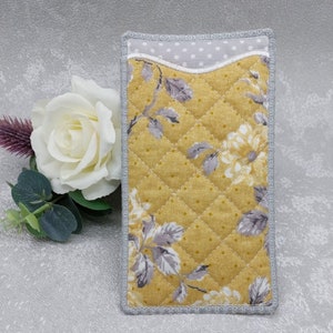May include: A rectangular quilted fabric case featuring a mustard yellow background with a floral pattern of grey leaves and white flowers, accented by a grey and white polka dot interior lining and finished with a grey stitched border trim.
