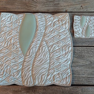 May include: A quilted placemat and coaster set. The placemat has a zebra print in beige and white, with a light green central panel. The coaster echoes the design. Both items have a quilted texture and a decorative edge.