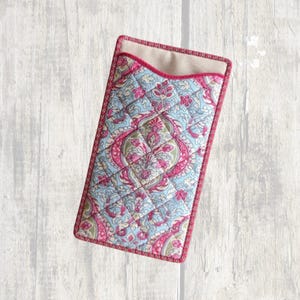 May include: A rectangular quilted fabric case featuring a light blue and pink paisley floral pattern. The case has a curved top opening with a solid pink fabric trim along the edges and a beige interior lining visible at the top opening.