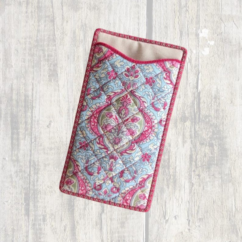 May include: A rectangular quilted fabric case featuring a light blue and pink paisley floral pattern. The case has a curved top opening with a solid pink fabric trim along the edges and a beige interior lining visible at the top opening.