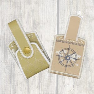 Puede incluir: Dos llaveros de cuero beige con costuras blancas. Un llavero tiene un diseño de brújula bordado.