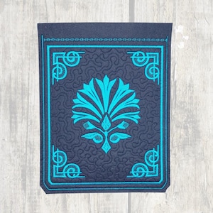 May include: A dark blue fabric patch with a teal blue embroidered design of a stylized flower with a decorative border.