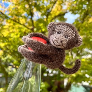 Könnte beinhalten: Eine kleine, handgestrickte braune Affenfigur mit einem hellbraunen Gesicht und Ohren. Der Affe ist aus flauschigem Garn gefertigt und so positioniert, als würde er den leuchtend roten Verschluss einer klaren Glasflasche mit seinem geschwungenen Schwanz umarmen.
