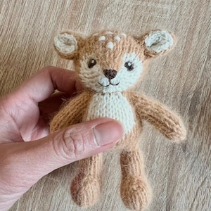 May include: A small, hand-knitted plush deer toy featuring a light brown body with a cream-coloured belly, face, and inner ears. The deer has embroidered dark brown eyes, a small nose, and a stitched mouth, with white spots on its forehead.