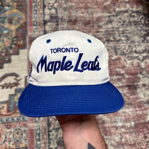 May include: A white and blue baseball cap featuring the text "TORONTO Maple Leafs" in blue script. The cap has a blue peak and a white crown. A sports accessory.