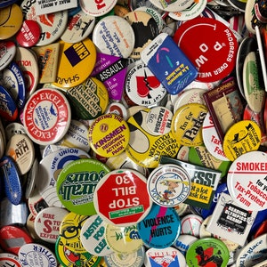 May include: A selection of vintage badges with diverse designs and text. The badges feature various colours, shapes, and messages, including political statements, event promotions, and humorous slogans. Some badges have text like "Violence Hurts" and "Stop Bill 30".