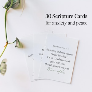 May include: A stack of white scripture cards with black text, including the words "30 Scripture Cards for anxiety and peace." One card displays a Bible verse from Deuteronomy 31:6. A green plant is in the background.