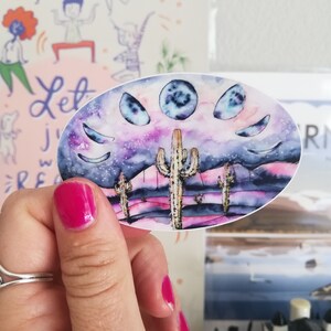 May include: An oval sticker featuring a watercolor painting of a desert landscape with cacti and a night sky with moon phases. The color palette includes purple, pink, and blue hues. The sticker is held by a hand with pink nail polish.