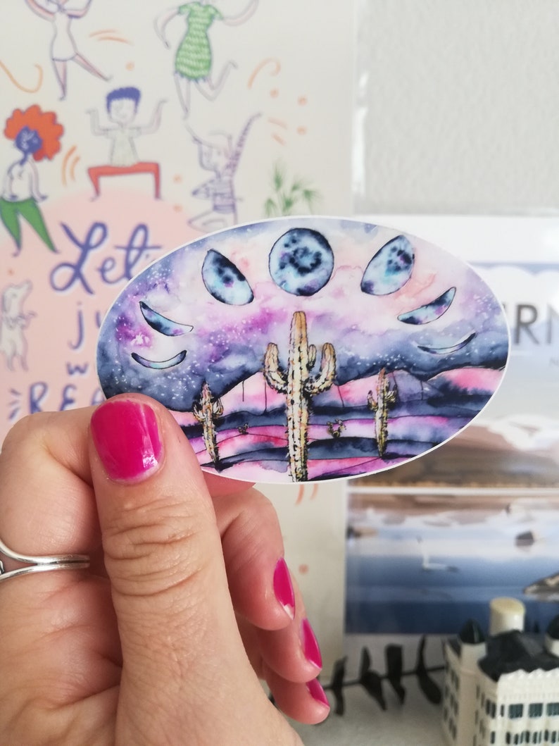 May include: An oval sticker featuring a watercolor painting of a desert landscape with cacti and a night sky with moon phases. The color palette includes purple, pink, and blue hues. The sticker is held by a hand with pink nail polish.