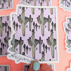 May include: A white sticker with a pink outline in the shape of the state of Arizona. The sticker has a pattern of cacti on a light purple background. The word "Arizona" is written in cursive at the bottom of the sticker.